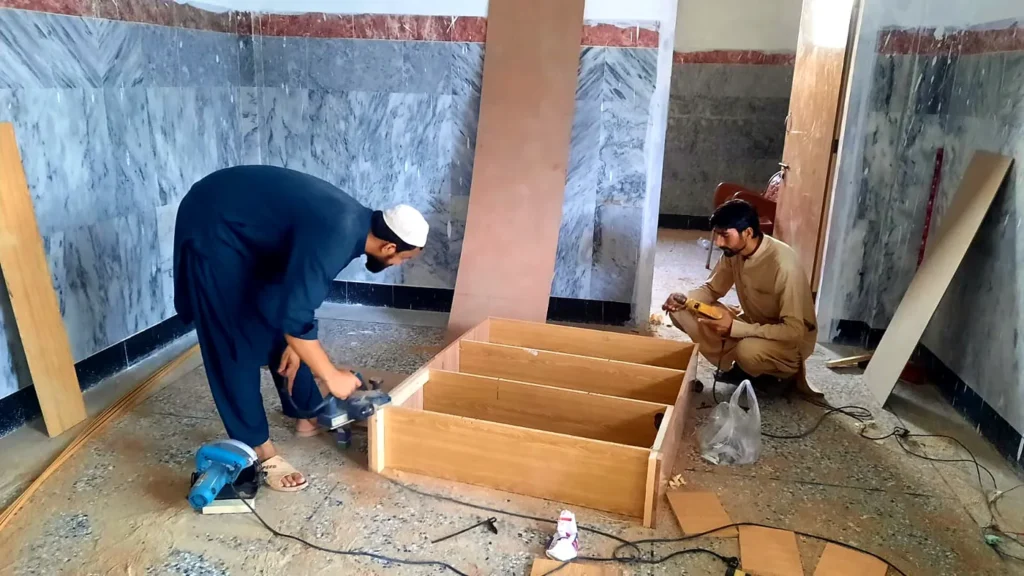 Two carpenters building a wooden cabinet on a classroom floor.