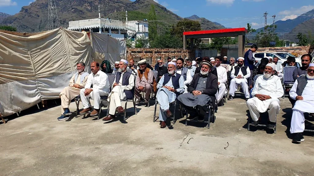 Elders and parents seated in rows at a school event with mountains behind.